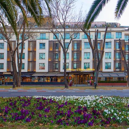 Dosso Dossi Hotels & Spa Downtown Istanbul Exterior photo The photo shows a building with a modern design, featuring a facade with large windows and a mix of materials. In the foreground, there are colorful flower beds with purple and white flowers. The scene is framed by tall trees, and the overall setting