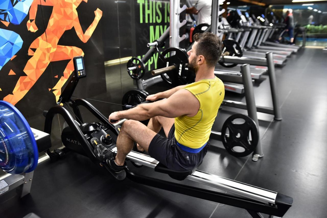 Dosso Dossi Hotels & Spa Downtown Istanbul Exterior photo The photo shows a man exercising on a rowing machine in a gym. He is wearing a yellow tank top and black shorts. The background features an exercise-themed mural with colorful graphics of a person in motion. Various gym equipment, including treadmill