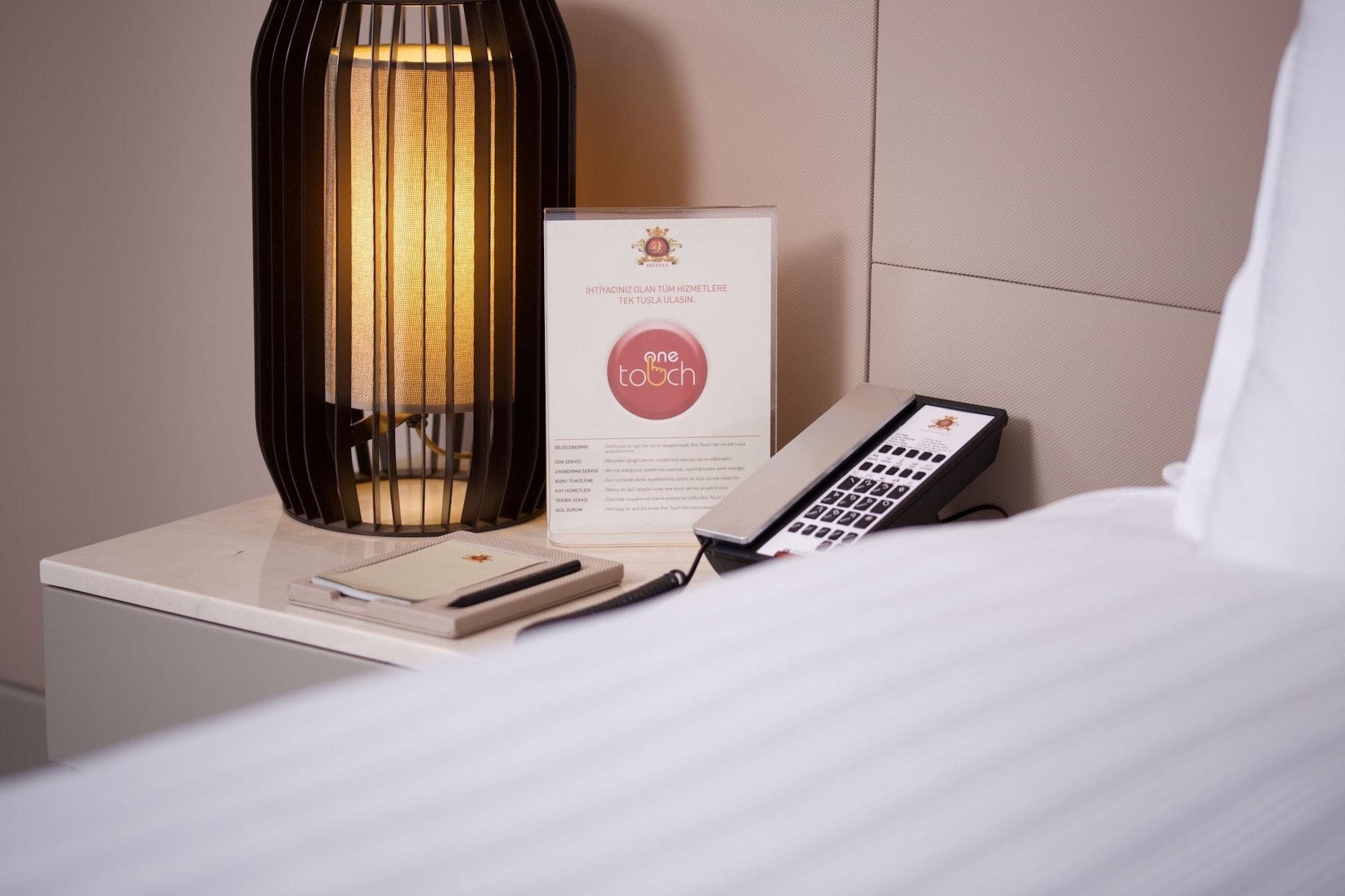 Dosso Dossi Hotels & Spa Downtown Istanbul Exterior photo The photo shows a bedside setup in a room. There is a stylish lamp with a warm light on a table, alongside a menu or information card that features a logo or brand name. A remote control is also placed on the table, suggesting it might be used for va