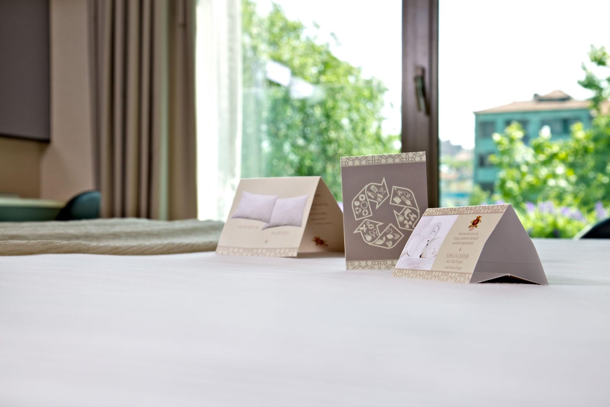Dosso Dossi Hotels & Spa Downtown Istanbul Exterior photo The photo shows a neatly made bed with a soft, white bedspread. In the foreground, there are three triangular cards placed upright on the bed. Each card has different designs or text, with one appearing to feature a pillow image and another possibly 