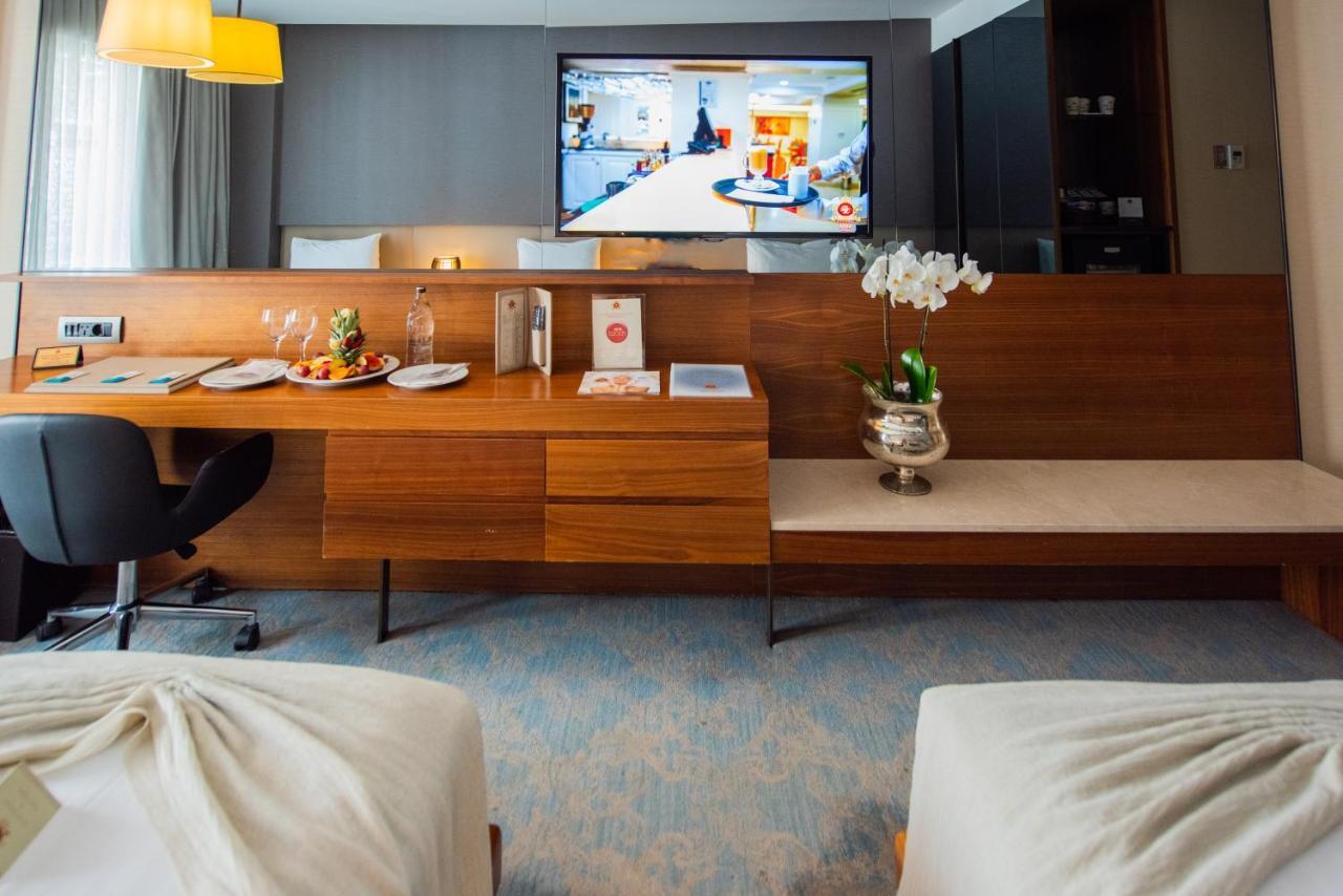 Dosso Dossi Hotels & Spa Downtown Istanbul Exterior photo The photo shows a modern hotel room featuring two beds with light-colored bedding. In the background, there is a flat-screen television mounted on the wall displaying what appears to be a hotel service or entertainment program. A wooden console table