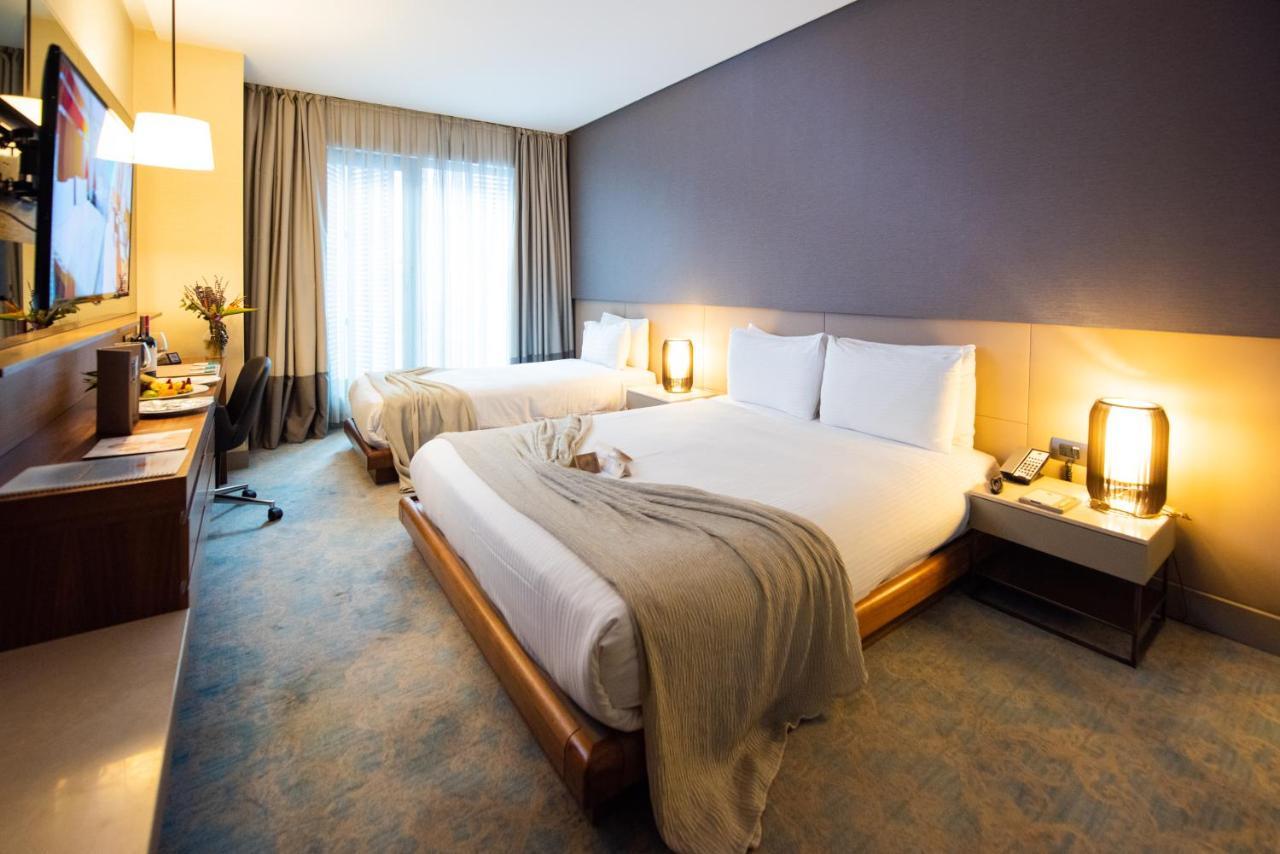 Dosso Dossi Hotels & Spa Downtown Istanbul Exterior photo The photo shows a modern hotel room with two beds. One bed is covered with a light-colored blanket, and there are white pillows on both beds. The walls are painted in a soft, neutral tone, while one wall is a darker shade. There are large windows wit