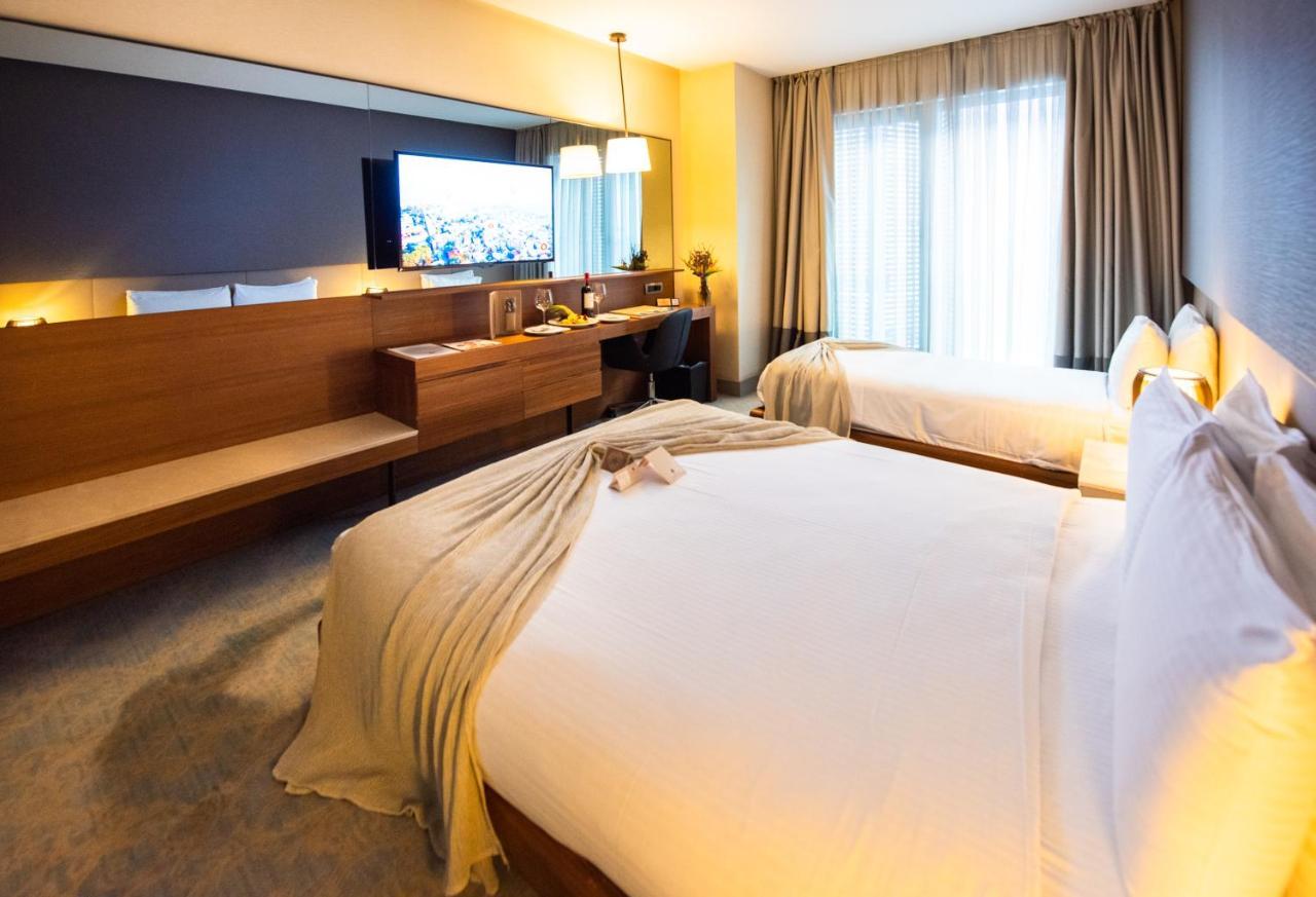 Dosso Dossi Hotels & Spa Downtown Istanbul Exterior photo The photo shows a modern hotel room featuring two beds with white linens and a decorative throw blanket. There's a wooden desk and a chair alongside a large flat-screen TV mounted on the wall. The room has large windows with curtains, allowing natura