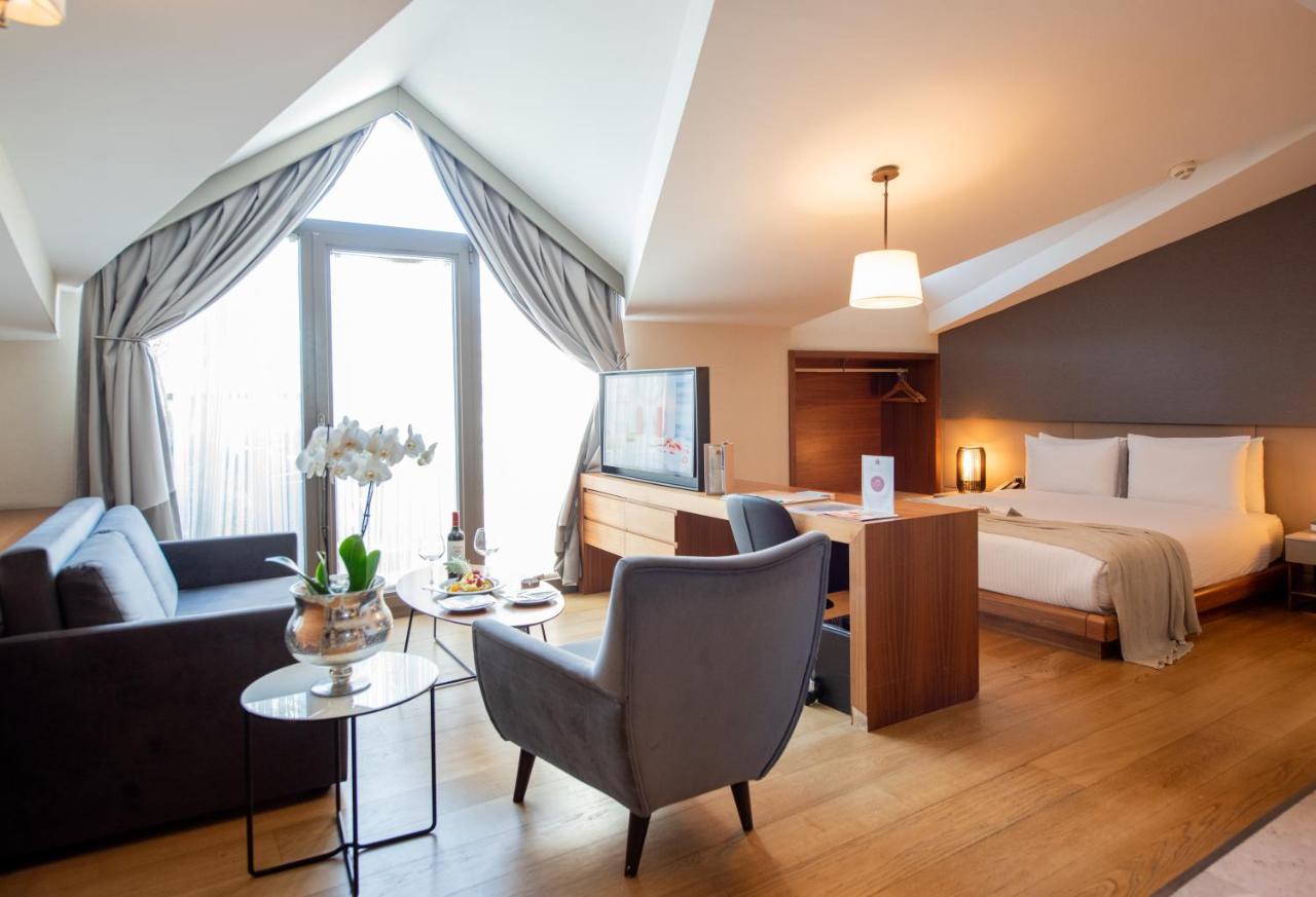 Dosso Dossi Hotels & Spa Downtown Istanbul Exterior photo The photo shows a stylish hotel room with a modern design. It features a comfortable bed in one area, complemented by a cozy seating area with a sofa and a chair. There is a small round table with a vase of flowers and some snacks. A flat-screen TV i