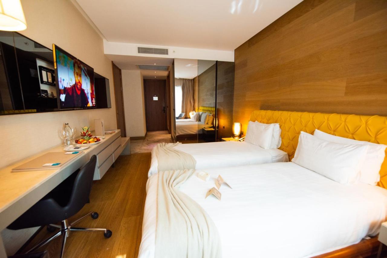 Dosso Dossi Hotels & Spa Downtown Istanbul Exterior photo The photo shows a modern hotel room featuring two single beds with white linens. There is a stylish yellow accent wall behind the beds. A flat-screen television is mounted on the wall opposite the beds, and a sleek desk and chair are positioned next 