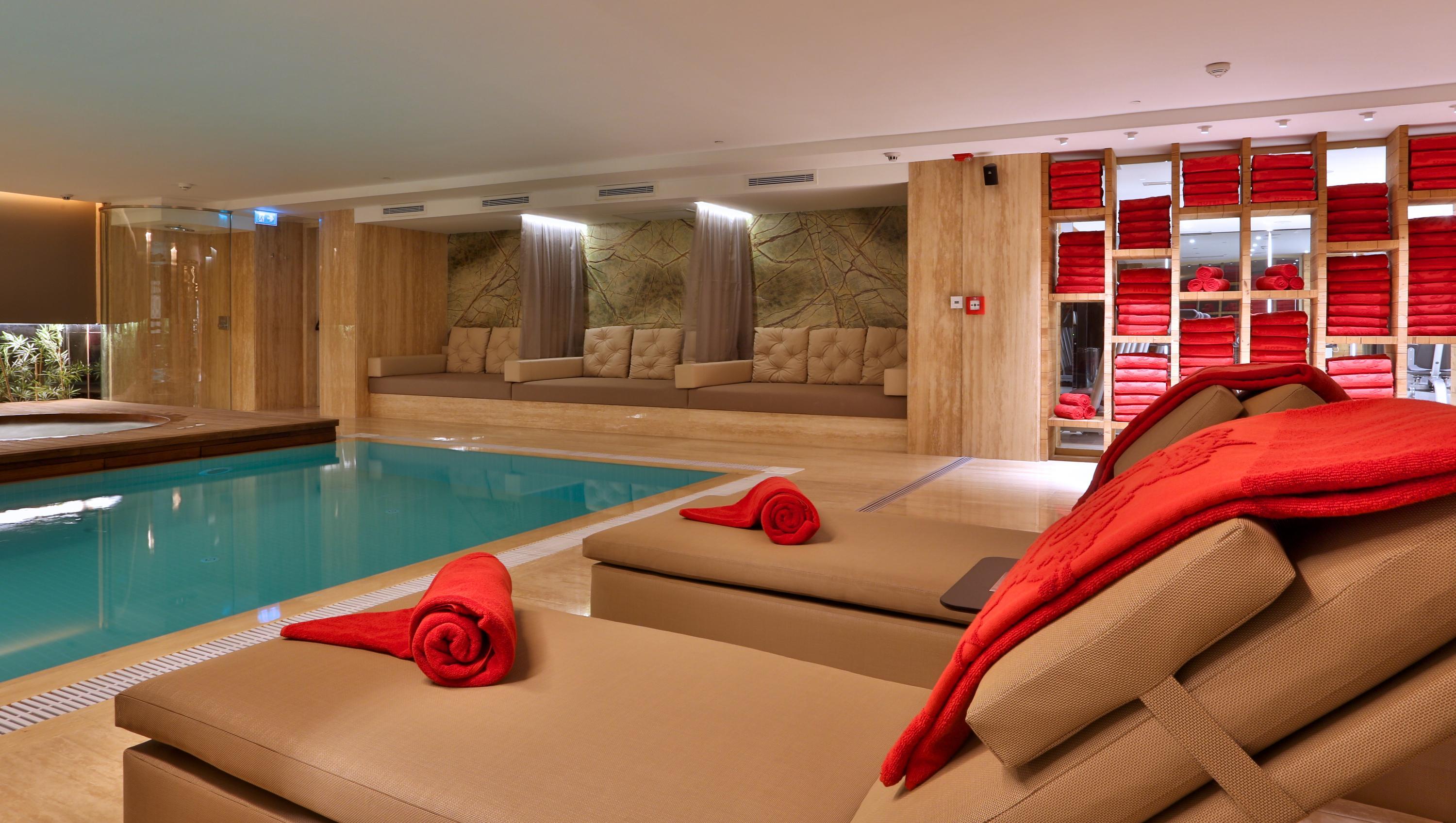 Dosso Dossi Hotels & Spa Downtown Istanbul Exterior photo The photo shows a modern, luxurious spa area featuring a swimming pool. In the foreground, there are comfortable lounge chairs with red towels rolled and placed on them. The walls are decorated with wood paneling, and there are several plush seating 