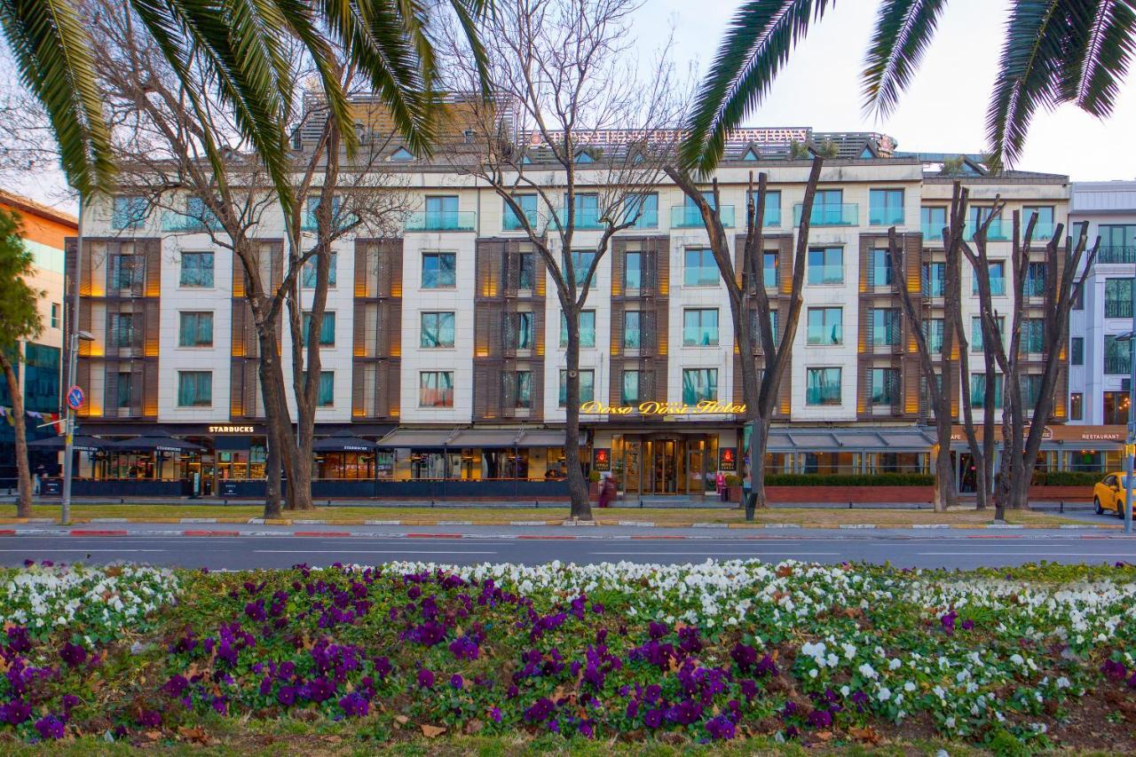 Dosso Dossi Hotels & Spa Downtown Istanbul Exterior photo The photo shows a building with a modern design, featuring a facade with large windows and a mix of materials. In the foreground, there are colorful flower beds with purple and white flowers. The scene is framed by tall trees, and the overall setting
