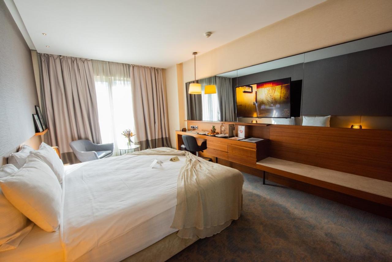 Dosso Dossi Hotels & Spa Downtown Istanbul Exterior photo The photo shows a modern hotel room featuring a large bed with white linens, set against a soft-toned wall. There is a stylish gray armchair in one corner and a sleek wooden desk with a chair. The room is well-lit, with large windows covered by light