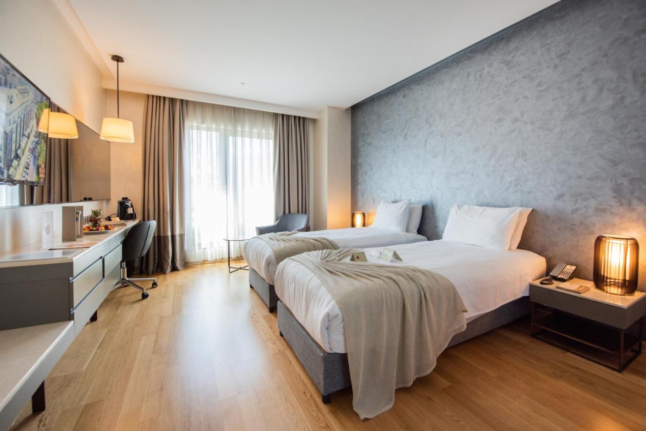 Dosso Dossi Hotels & Spa Downtown Istanbul Exterior photo The photo shows a modern hotel room featuring two single beds with white linens. The room has a muted color palette, including a gray wall and light wooden flooring. Large windows allow natural light to flood the space, and there are sheer curtains f