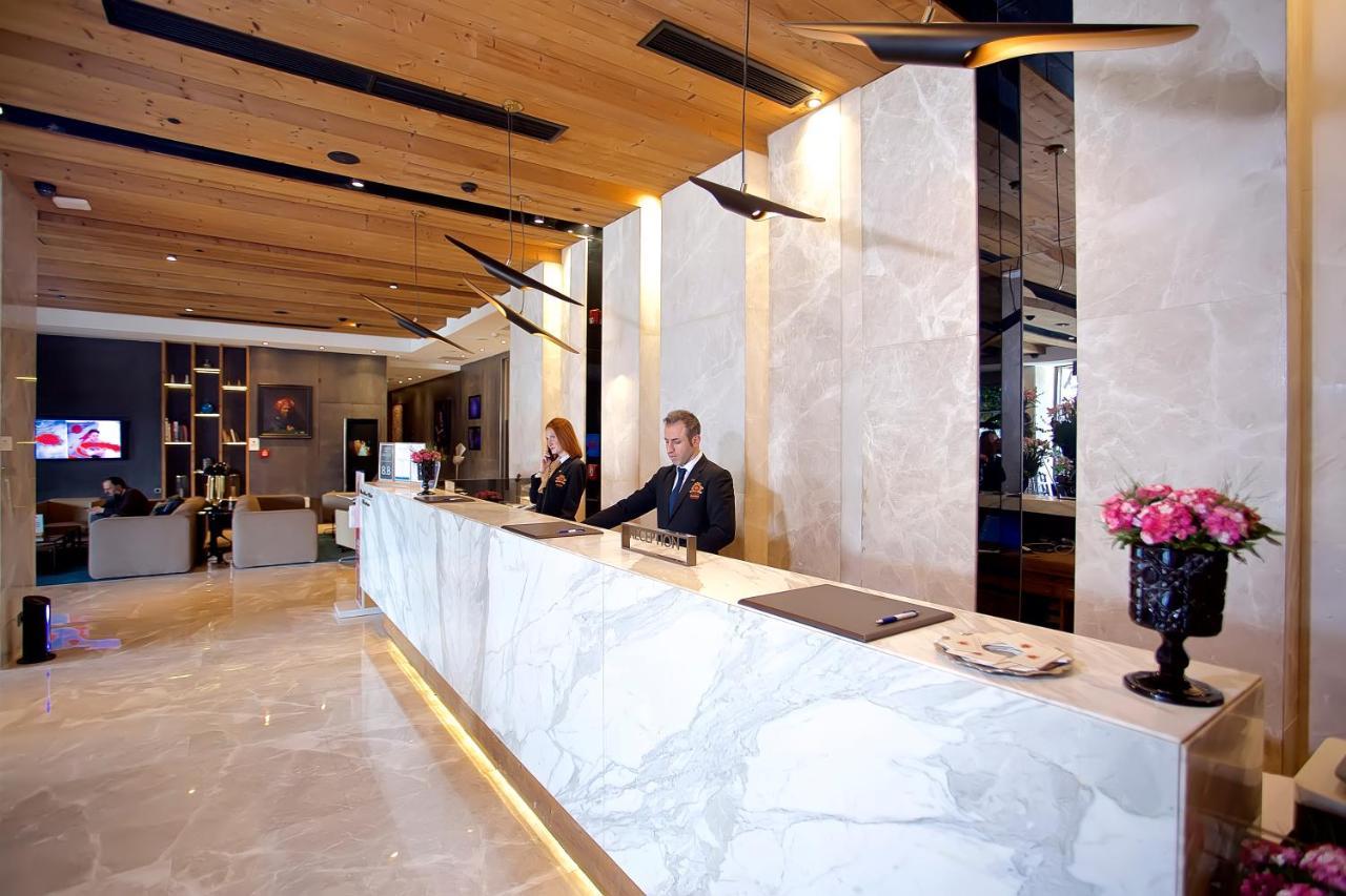 Dosso Dossi Hotels & Spa Downtown Istanbul Exterior photo The photo shows a hotel lobby reception area. The reception desk is elegantly designed with a marble surface and is situated against a backdrop of light-colored walls. There are two staff members behind the desk—one is a man in a suit, and the other 