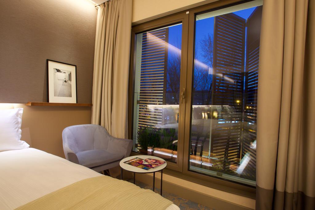Dosso Dossi Hotels & Spa Downtown Istanbul Exterior photo The photo depicts a modern hotel room. On the left side, there is a neatly made bed with a light-colored blanket. Next to the bed, there's a small round table with some magazines on top. A cozy chair in a light shade is positioned nearby. The main fe